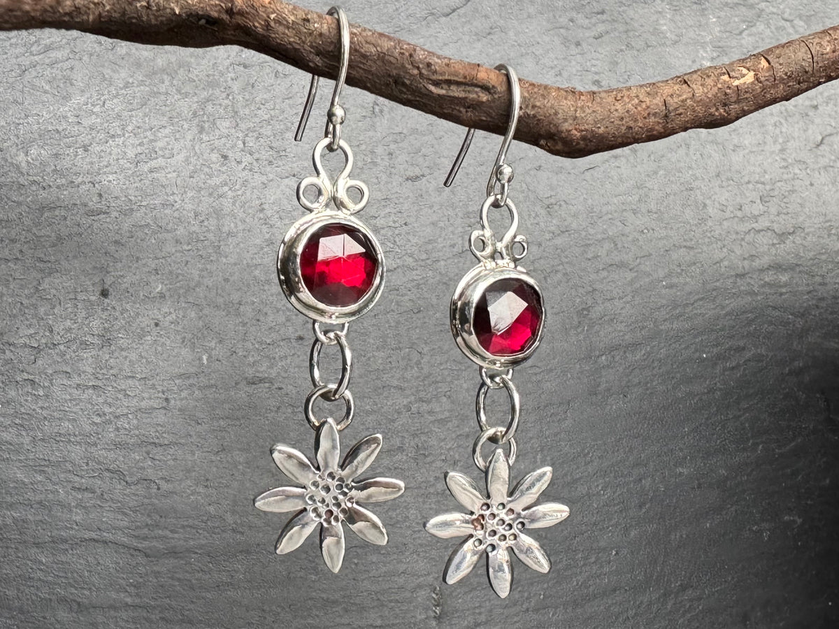 Silver earrings with red garnet gemstones and daisy flowers on a gray stone background.