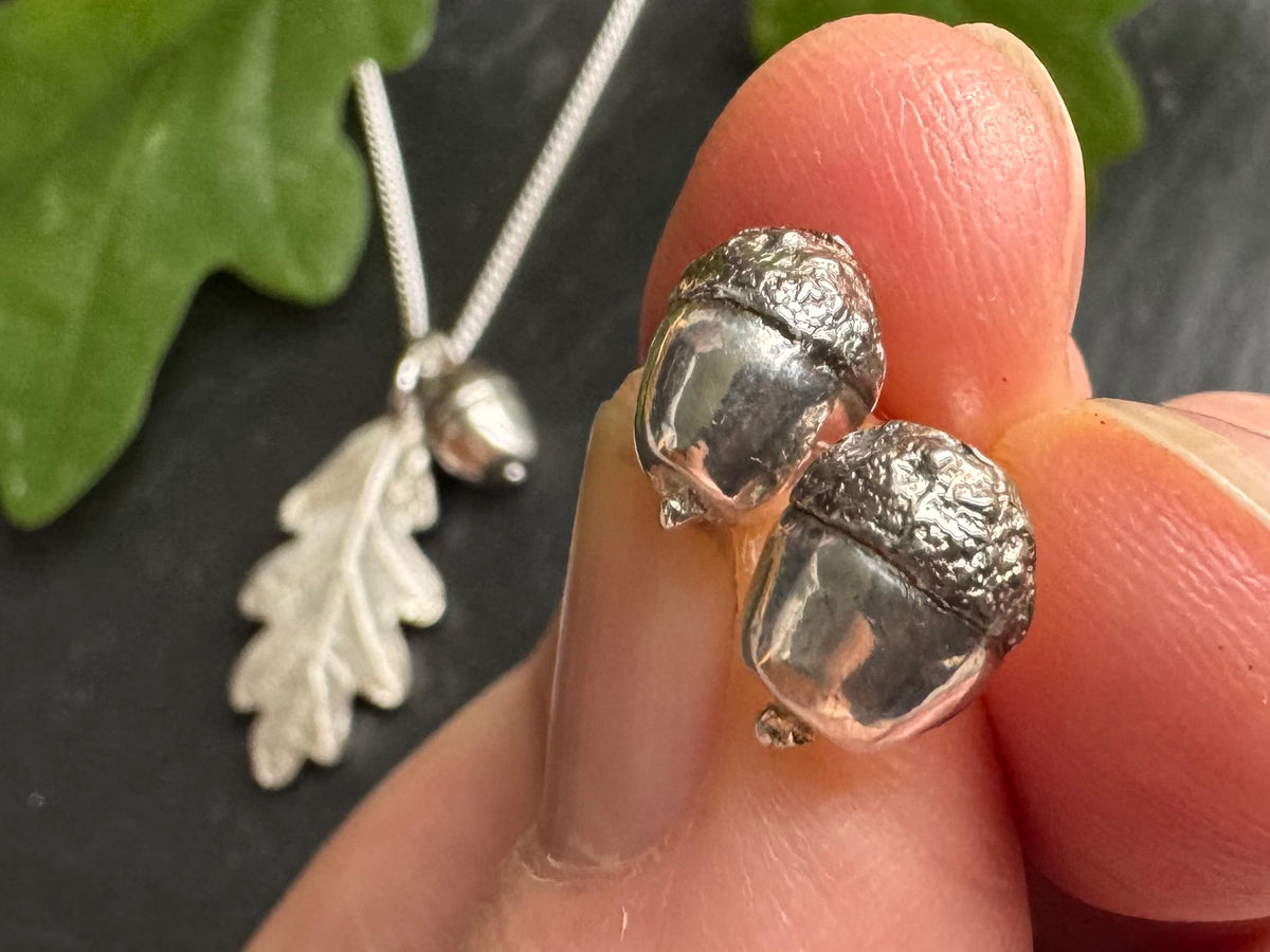 Silver acorn-shaped earrings held between fingers with a leaf pendant in the background.