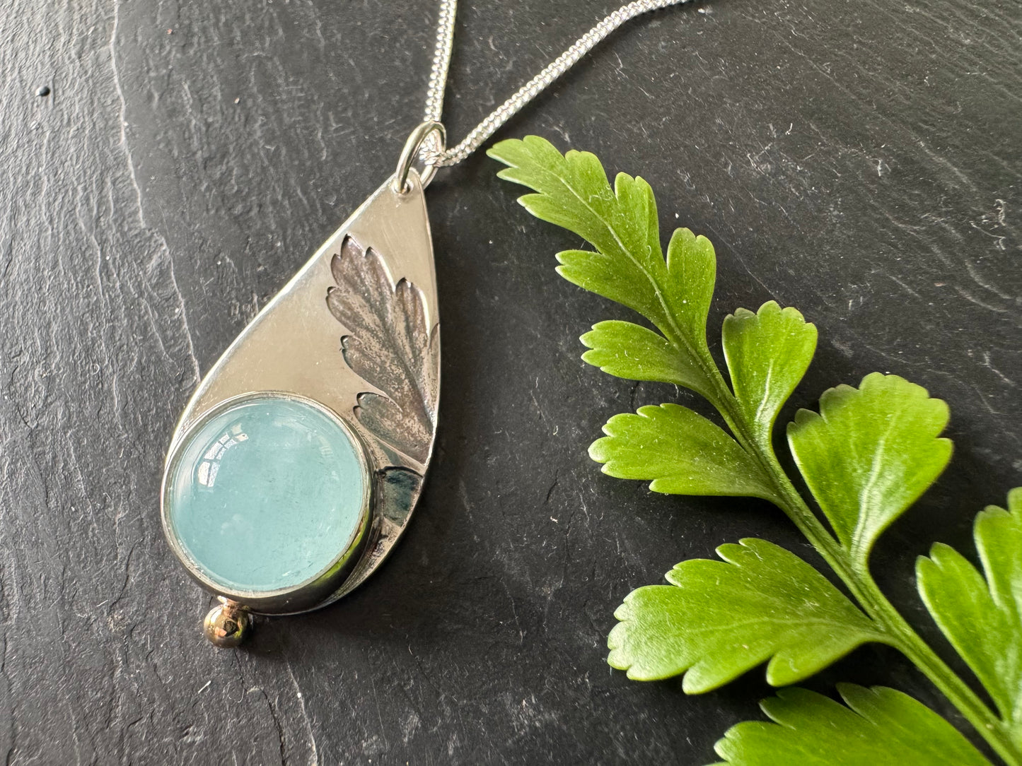 Silver pendant with anaquamarine stone on a dark surface with green leaves