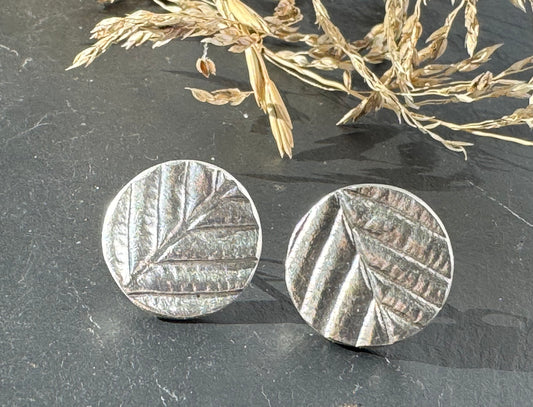Silver circular earrings with leaf pattern on a dark surface with dried grass.