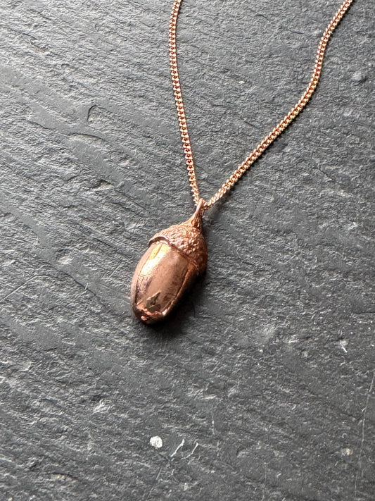 A copper acorn pendant necklace on a delicate chain, displayed on a dark textured slate surface.