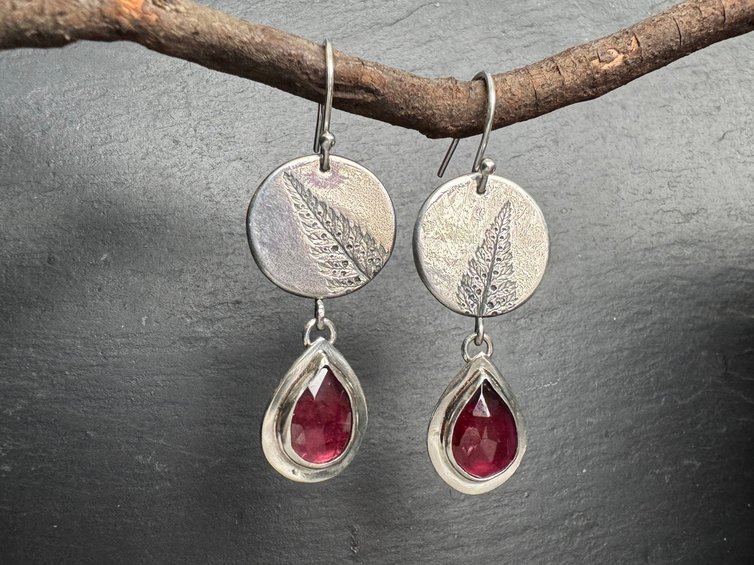 Silver earrings with red garnet teardrop stones hanging from a branch on a dark surface