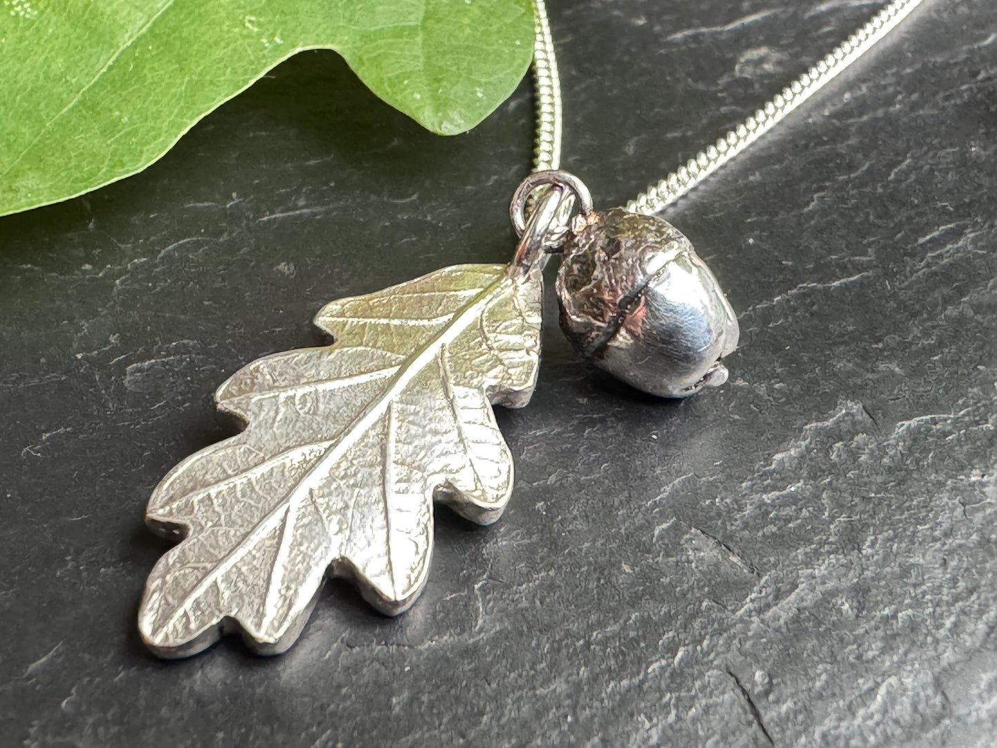 Silver leaf and acorn pendant on a chain against a dark surface with a green leaf.