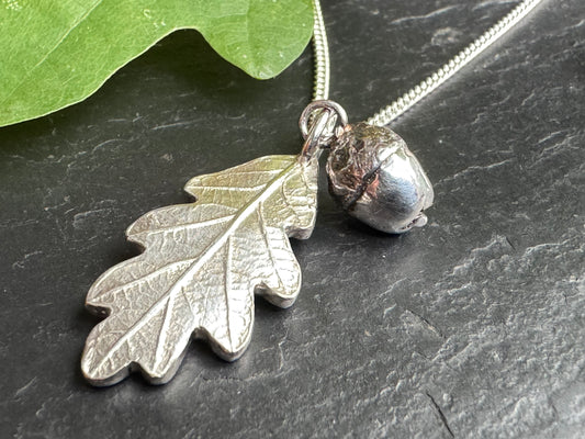 Silver leaf and acorn pendant on a chain against a dark surface with a green leaf.