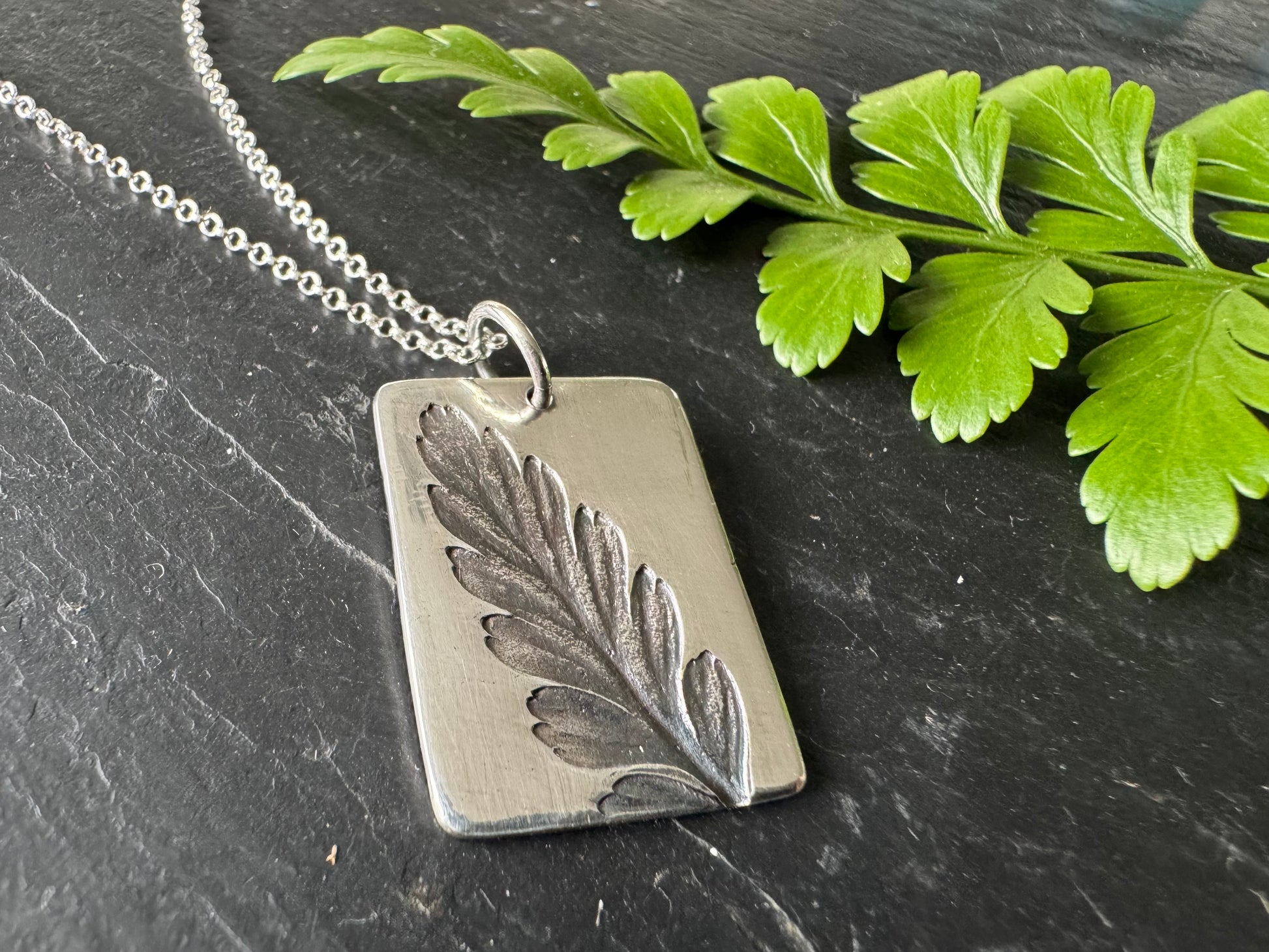 Silver pendant with leaf design on a chain, lying on a dark surface with green leaves.